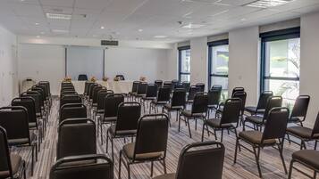Sala de conferência com fileiras de cadeiras, uma tela de projetor e uma mesa com arranjos florais.