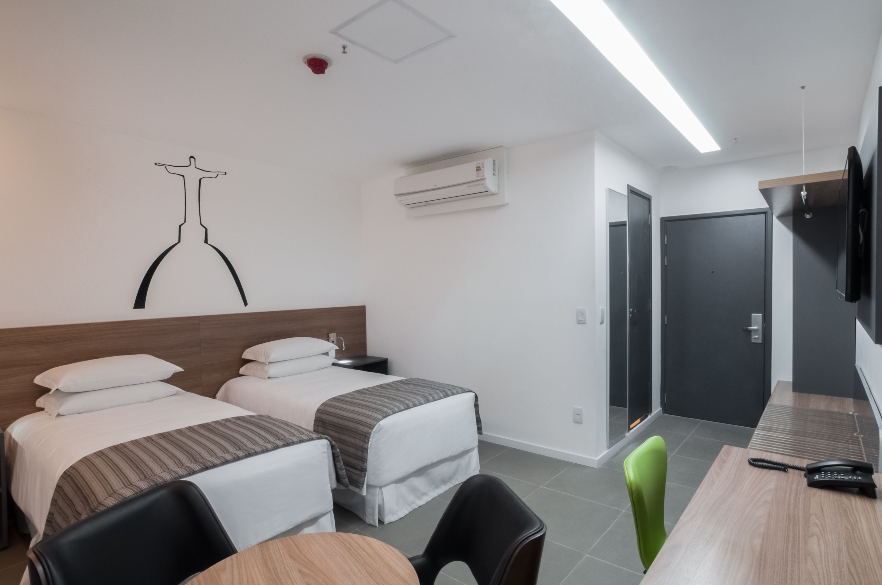 A hotel room with two beds, a desk, a chair, a TV, and an air conditioning unit.