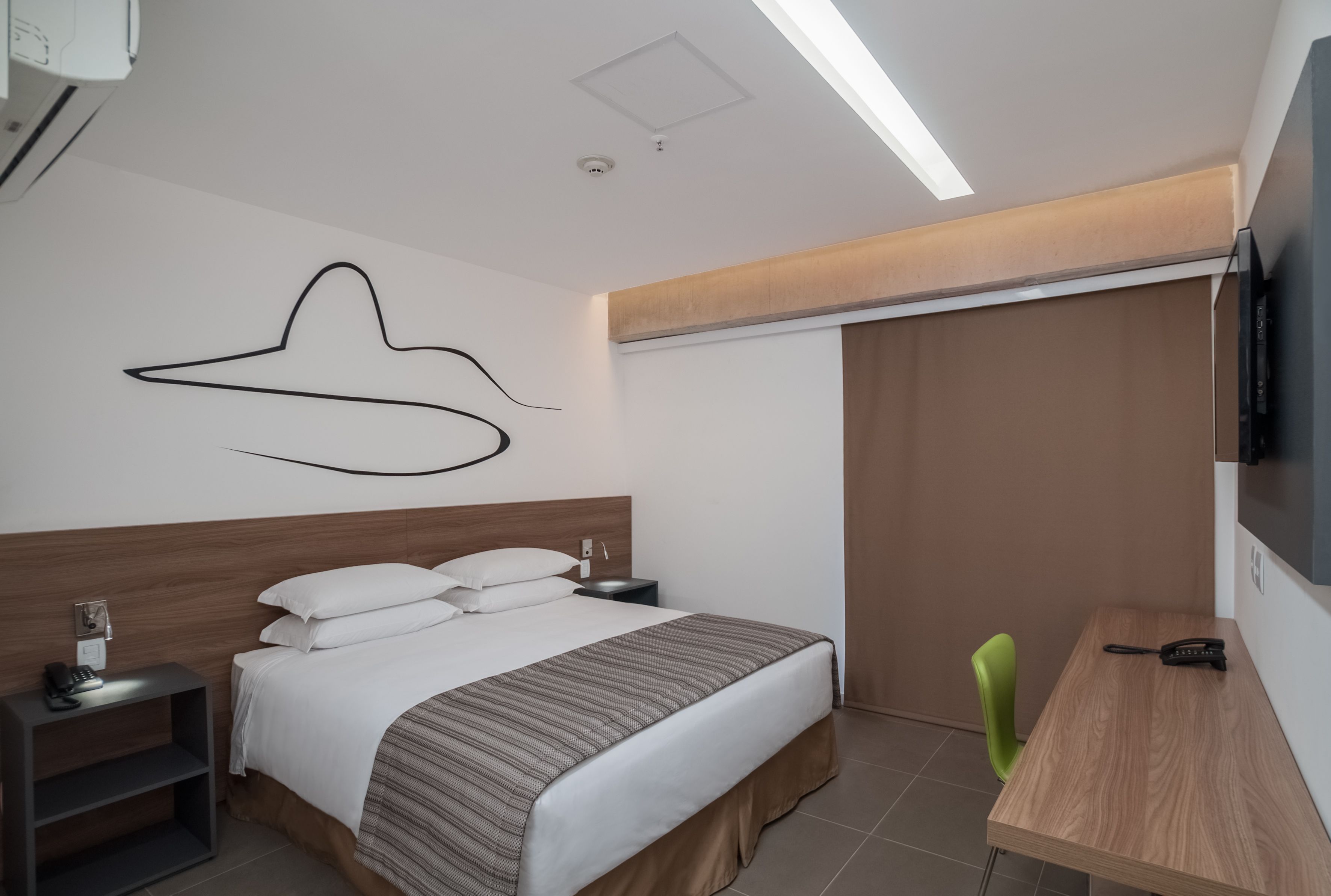 A modern hotel room with a large bed, bedside table, and a desk with a chair.