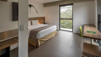 A modern hotel room with a large bed, a desk, and a window with a view of greenery.