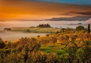 A panoramic view of a hilly landscape with a mist-covered valley and a small cluster of buildings on a hill. - Agriturismo La Buca di Bellugi (San Quirico d'Orcia)