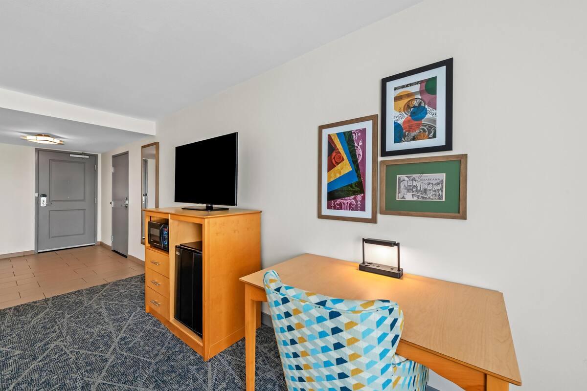 A hotel room with a desk, a flat-screen TV, a microwave, a small refrigerator, and a patterned chair.