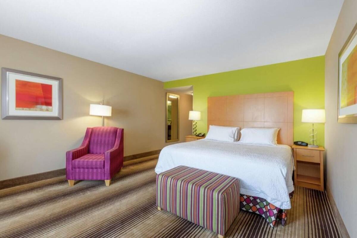A hotel room with a bed, a chair, a nightstand, a lamp, a framed picture, and a striped bench.