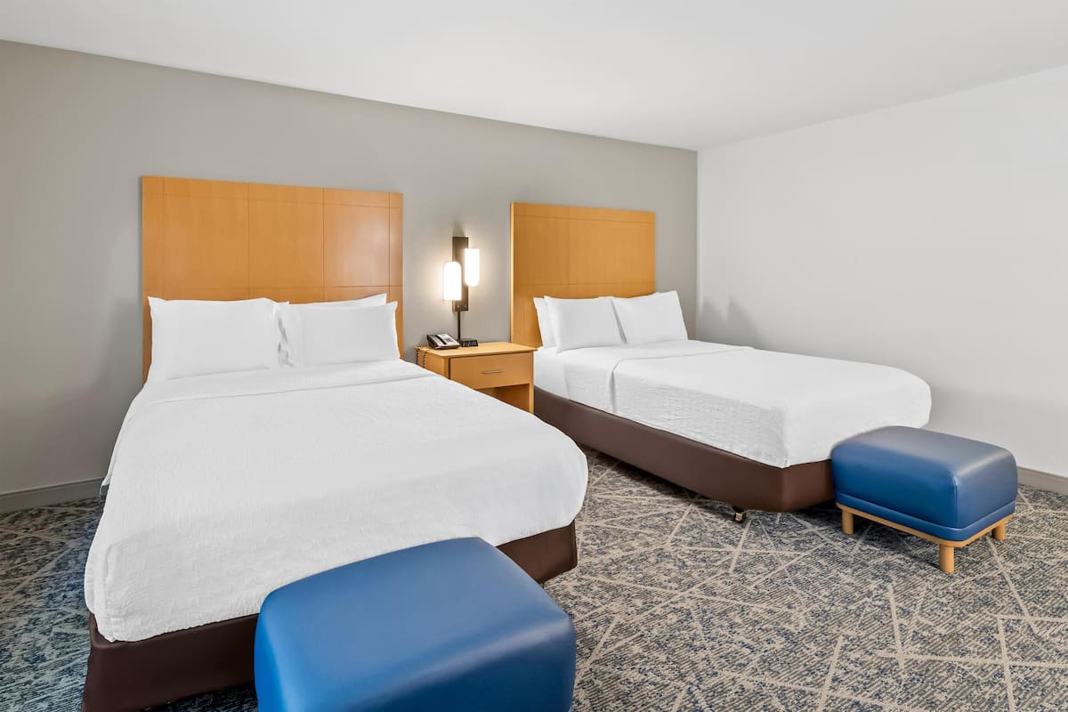 A hotel room with two beds, a nightstand, a lamp, and a blue ottoman.