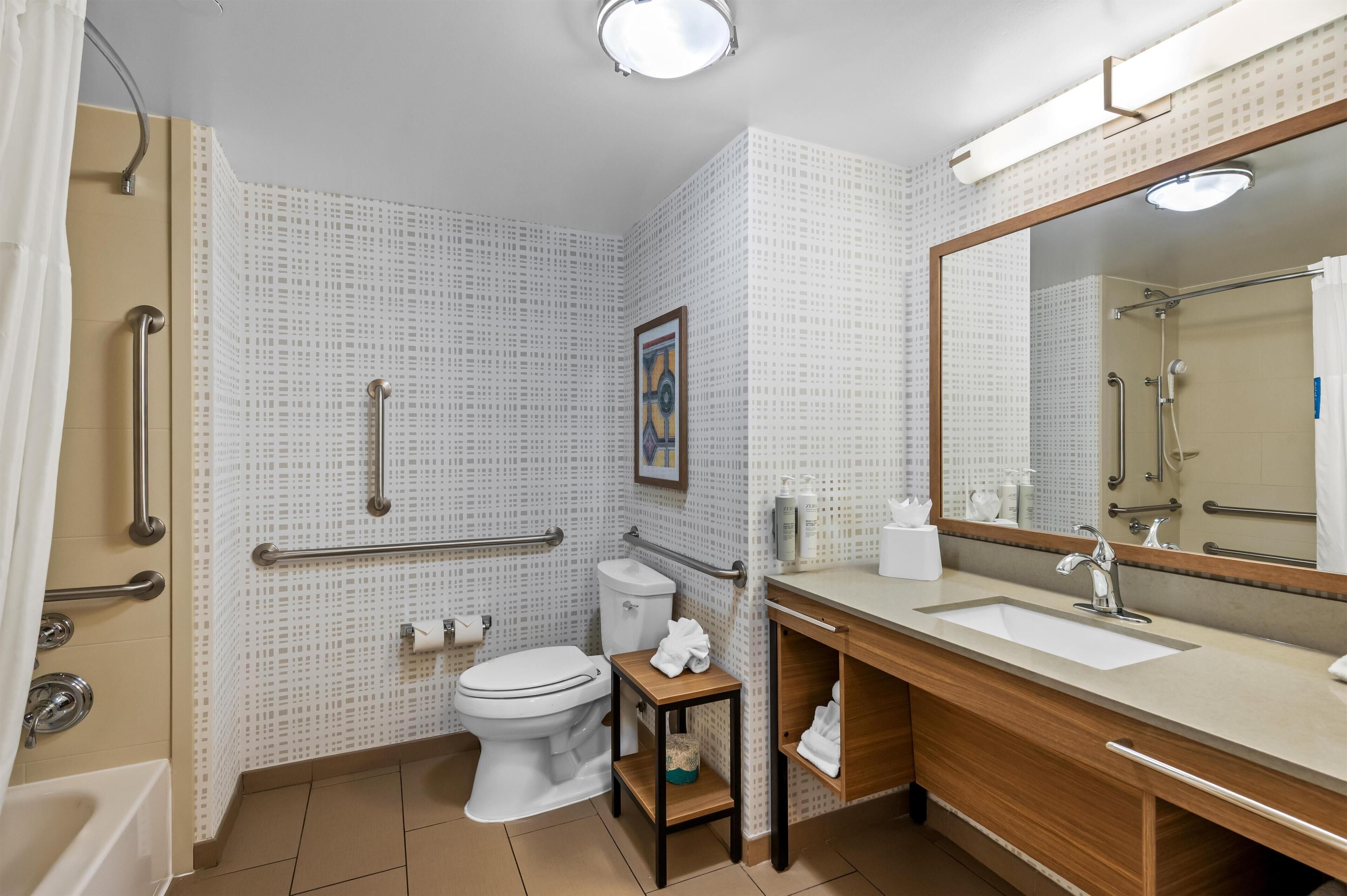 Un baño con inodoro, lavamanos y ducha. Cuenta con barras de apoyo y ducha accesible para silla de ruedas.