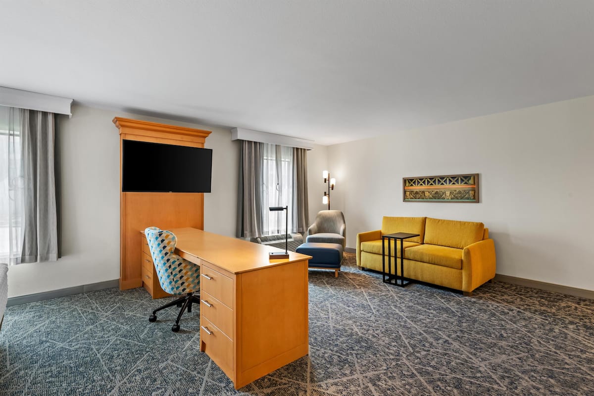 A hotel room with a wooden desk, a yellow sofa, a patterned carpet, and a painting on the wall.