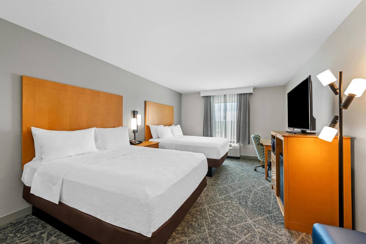 A hotel room with two beds, a desk, a television, and a desk lamp.