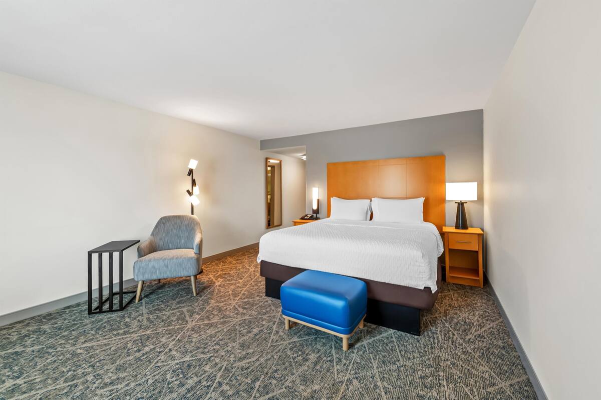 A hotel room with a large bed, a chair, a small table, and a nightstand with a lamp.