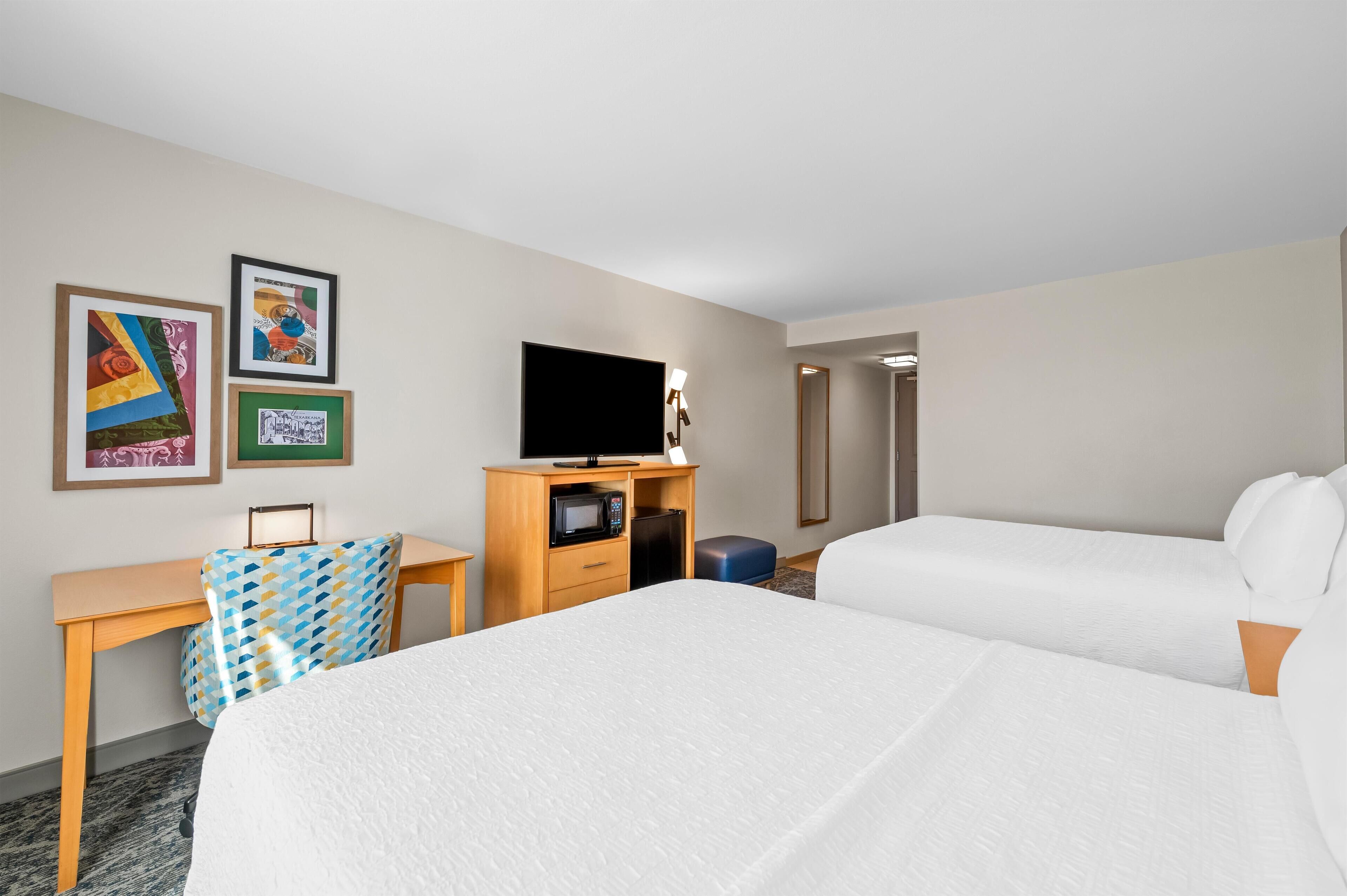 A hotel room with two beds, a desk with a TV, a microwave, and artwork on the wall.