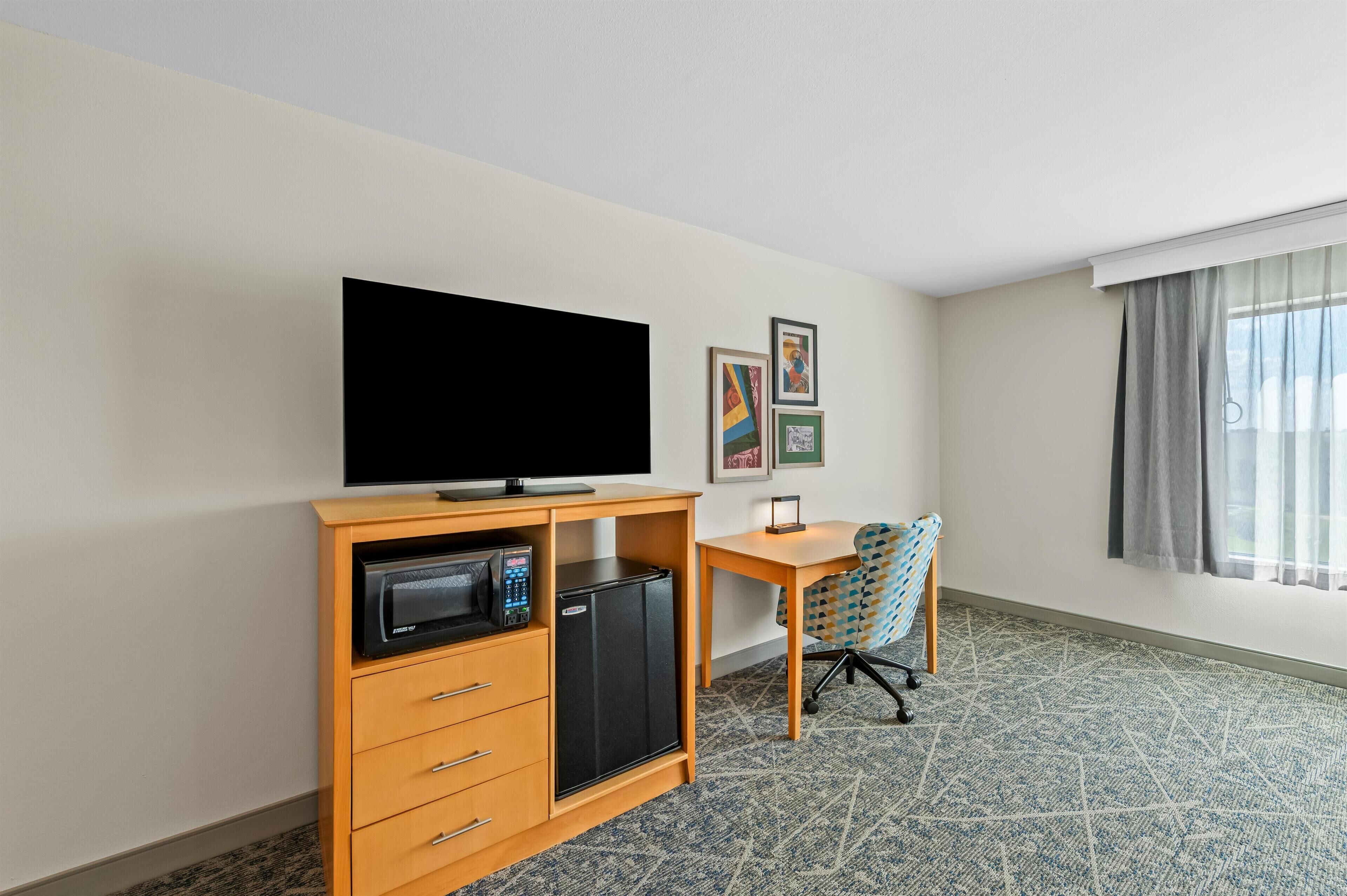 A hotel room with a flat-screen TV, a microwave, a refrigerator, a desk, a patterned carpet, and a window with curtains.
