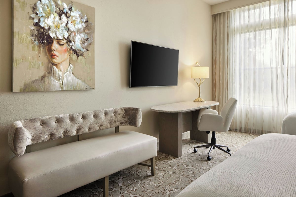 A hotel room with a bed, a sofa, a desk, a chair, a TV, and a painting on the wall.