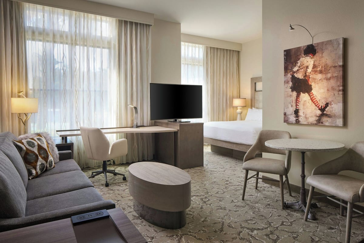 A hotel room with a sofa, a desk with a flat-screen TV, a bed, two armchairs, a small round table, and a painting on the wall.