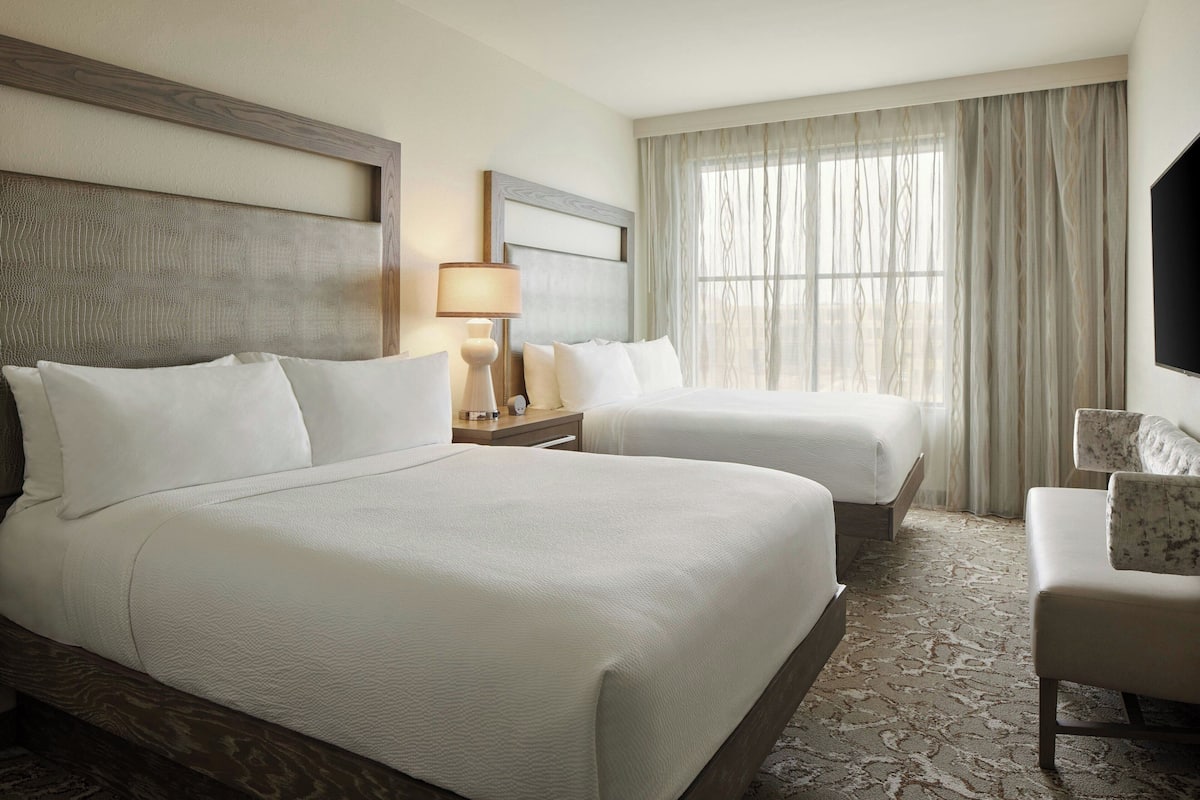 A hotel room with two beds, a flat-screen TV, a desk with a lamp, and a window with curtains.