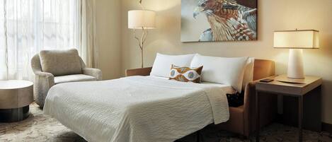 A hotel room with a bed, a chair, a lamp, a painting, and a carpet.