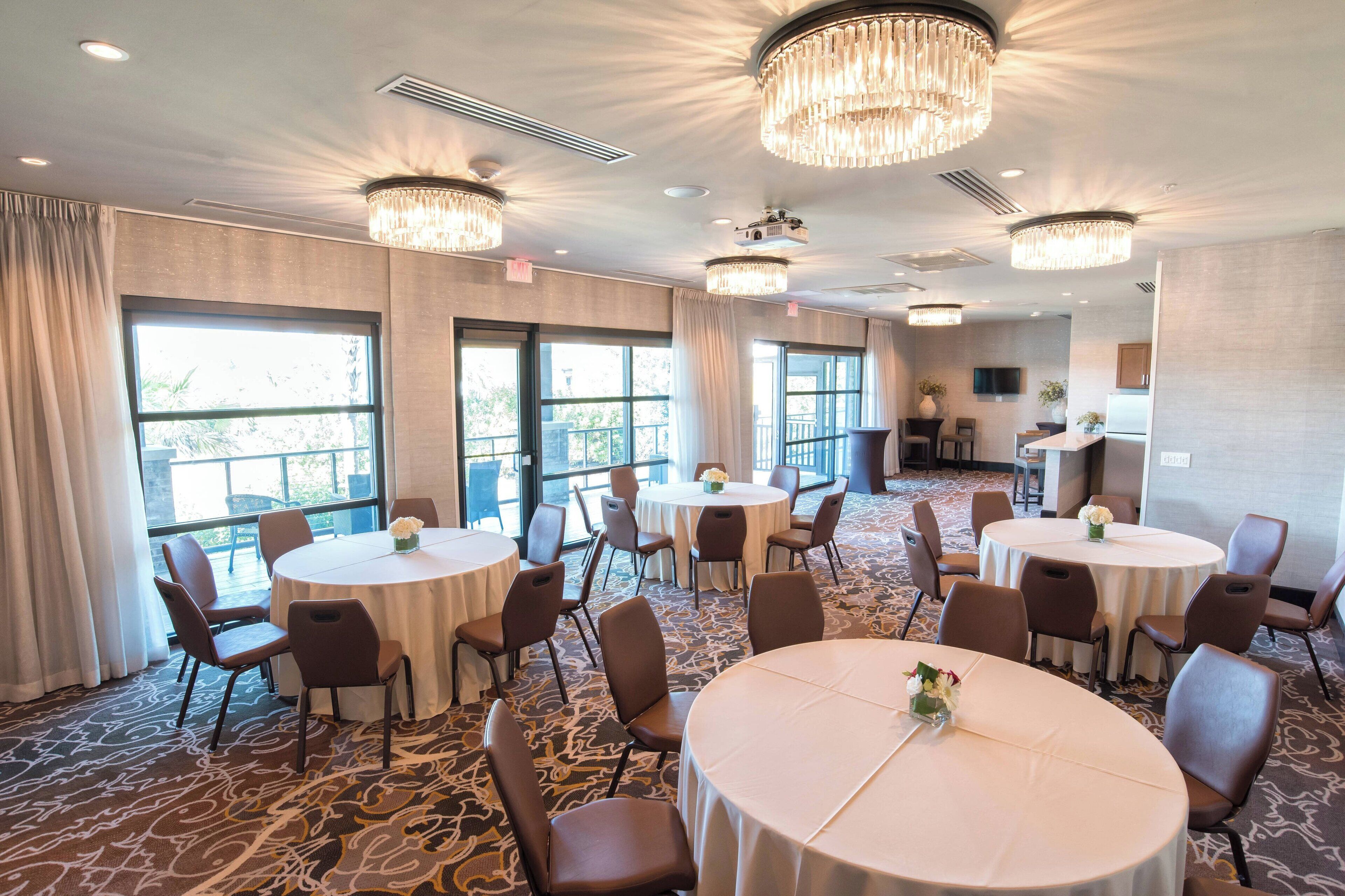A hotel conference room with multiple round tables, chairs, and large windows.