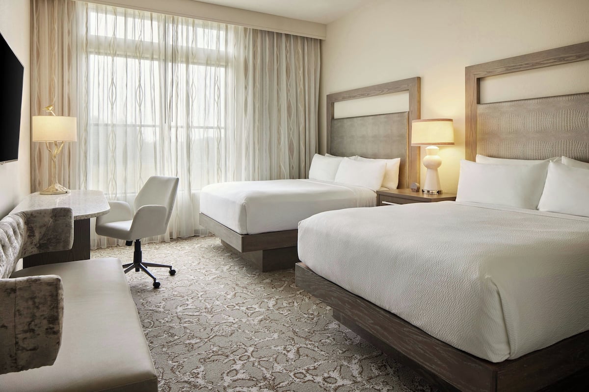 A hotel room with two beds, a desk, a chair, and a large window with curtains.