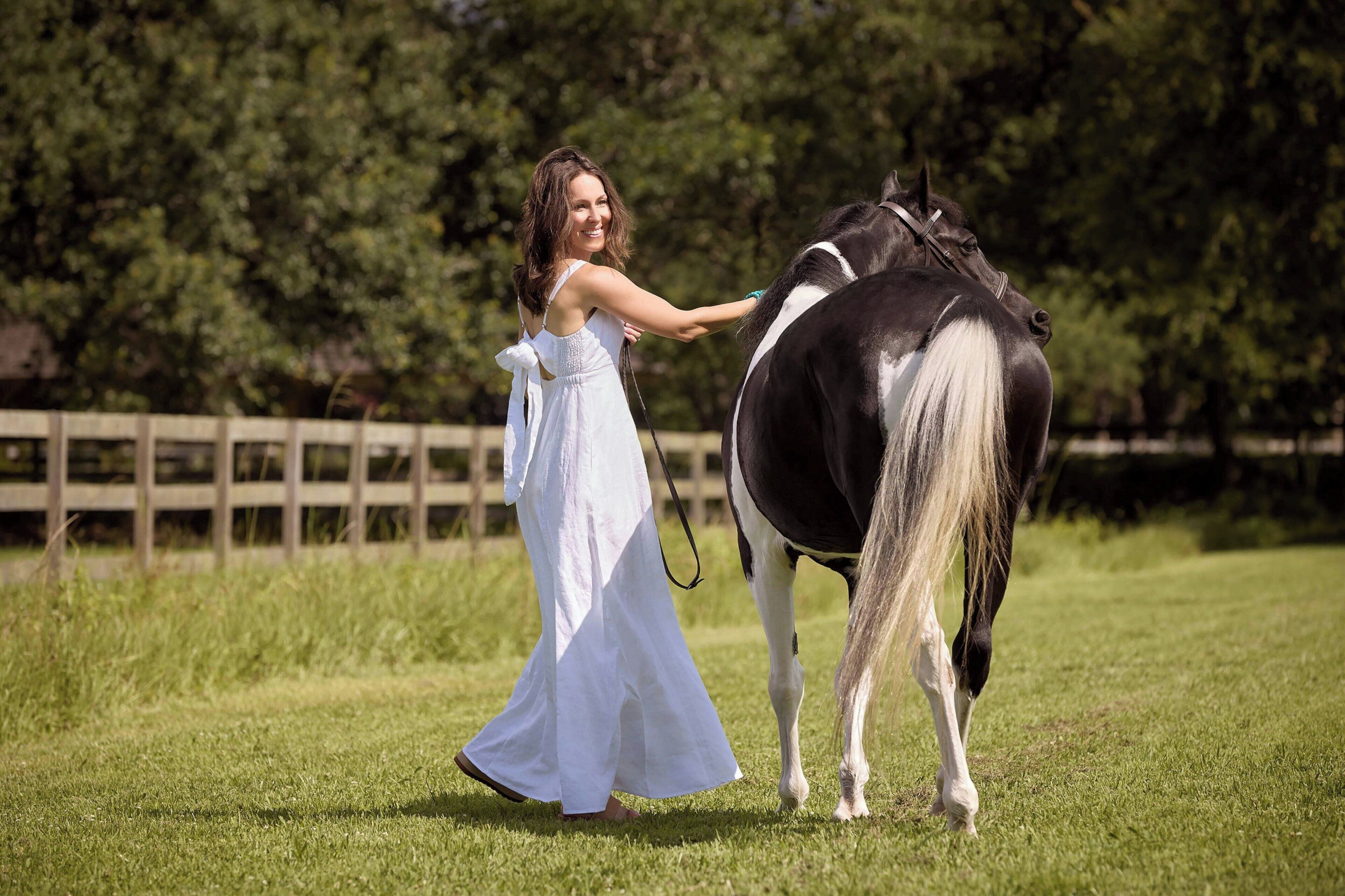 Una donna con un abito bianco conduce un cavallo bianco e nero in un prato, con una recinzione di legno sullo sfondo.
