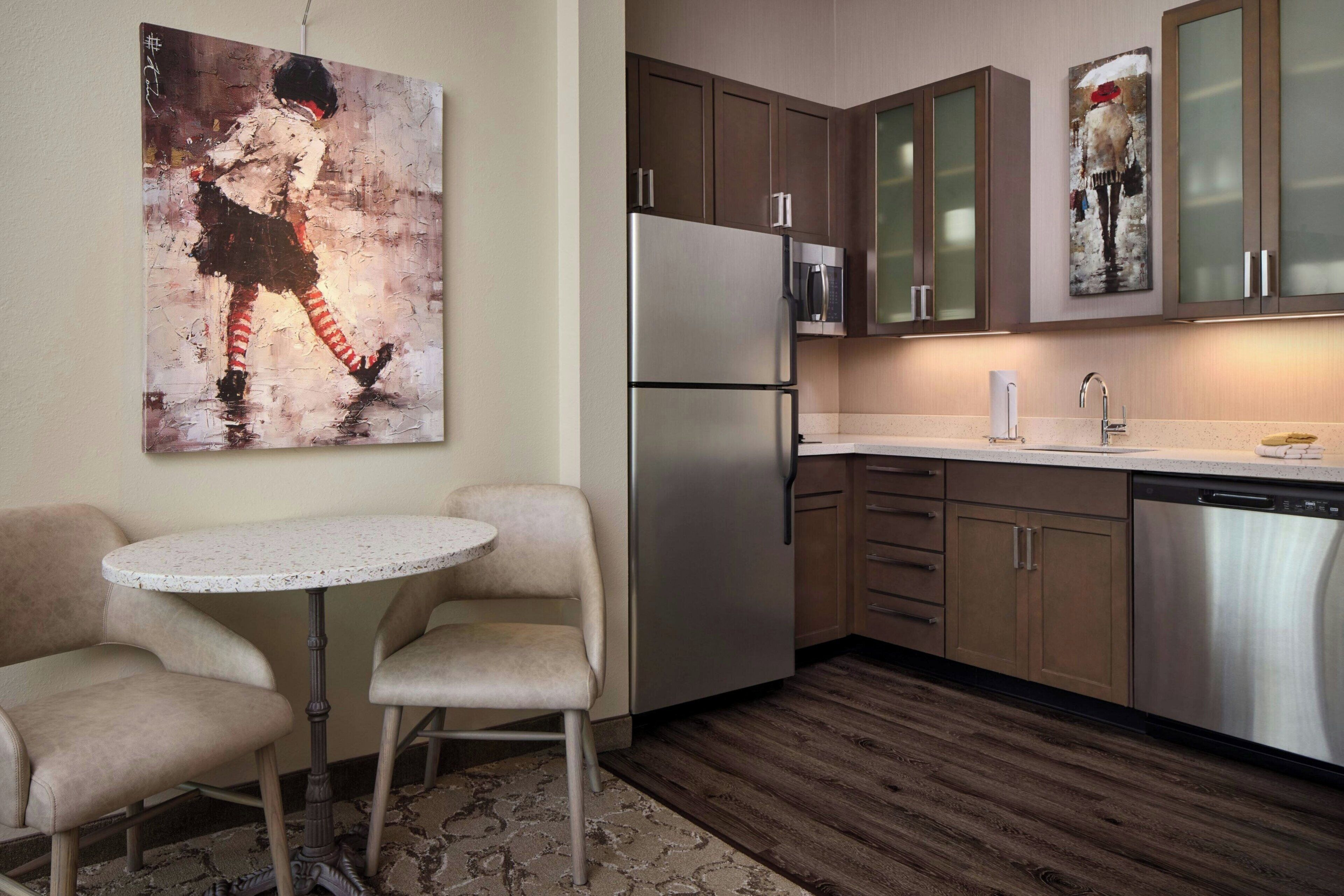 A modern kitchen with a stainless steel refrigerator, microwave, and dishwasher, wooden cabinets, and a round table with two chairs.