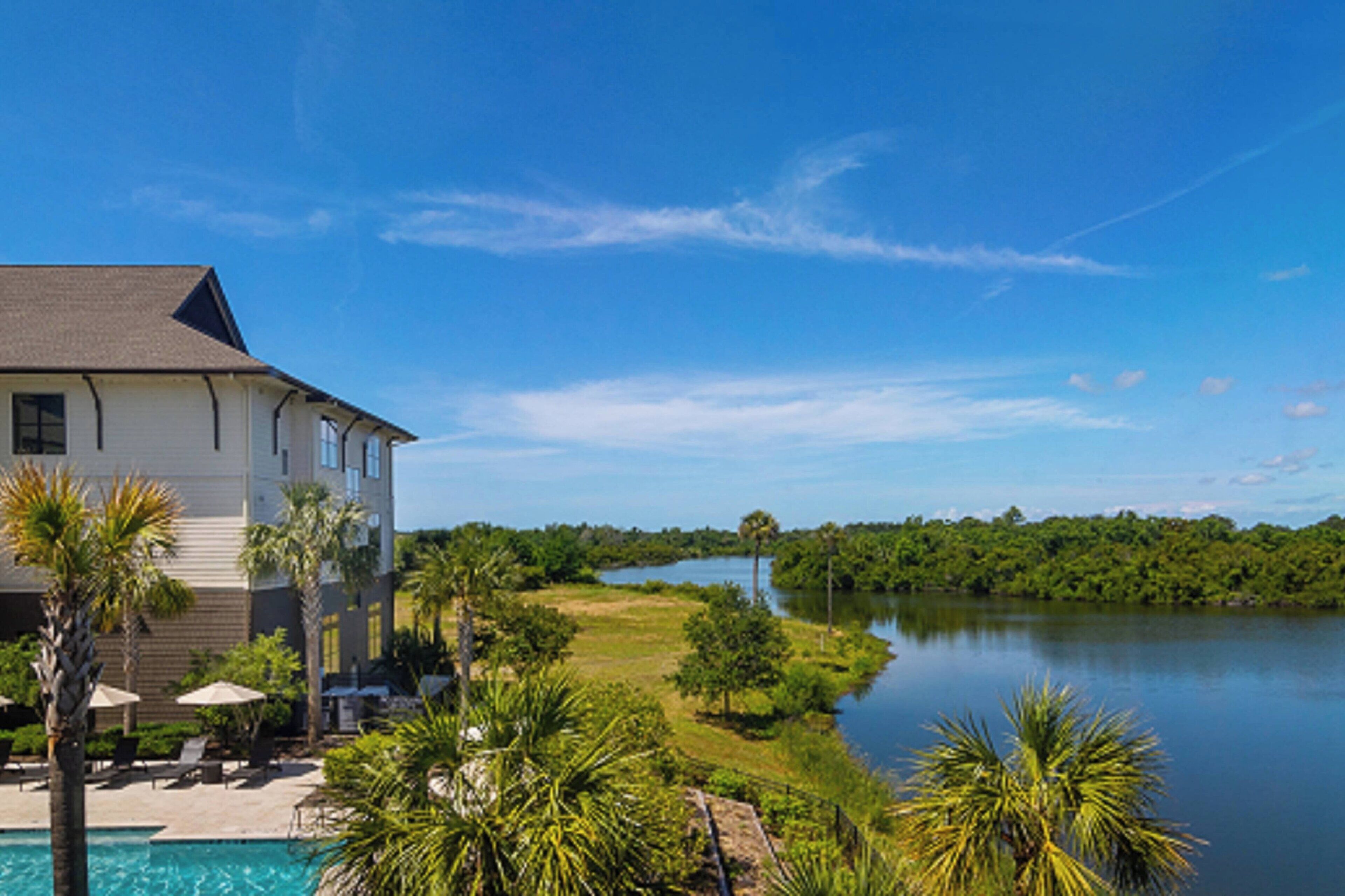 A large building with a swimming pool and palm trees, overlooking a calm lake and dense greenery.