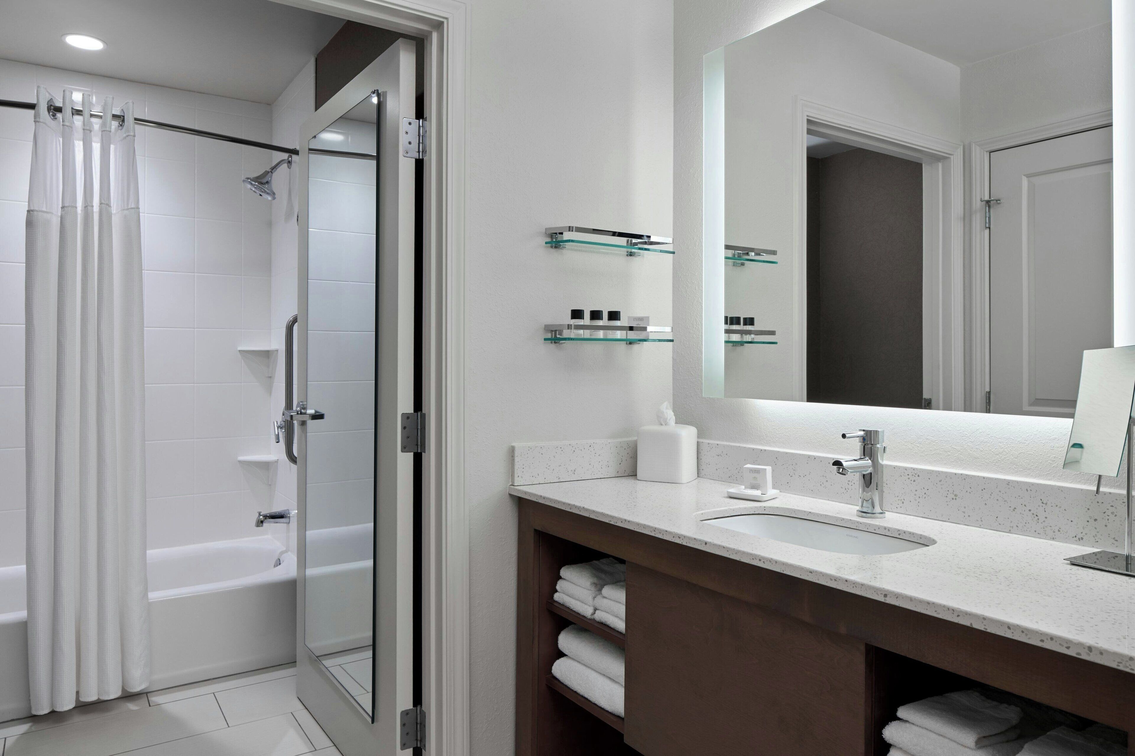 A modern bathroom with a glass shower enclosure, a bathtub, a sink with a mirror, and glass shelves with toiletries.