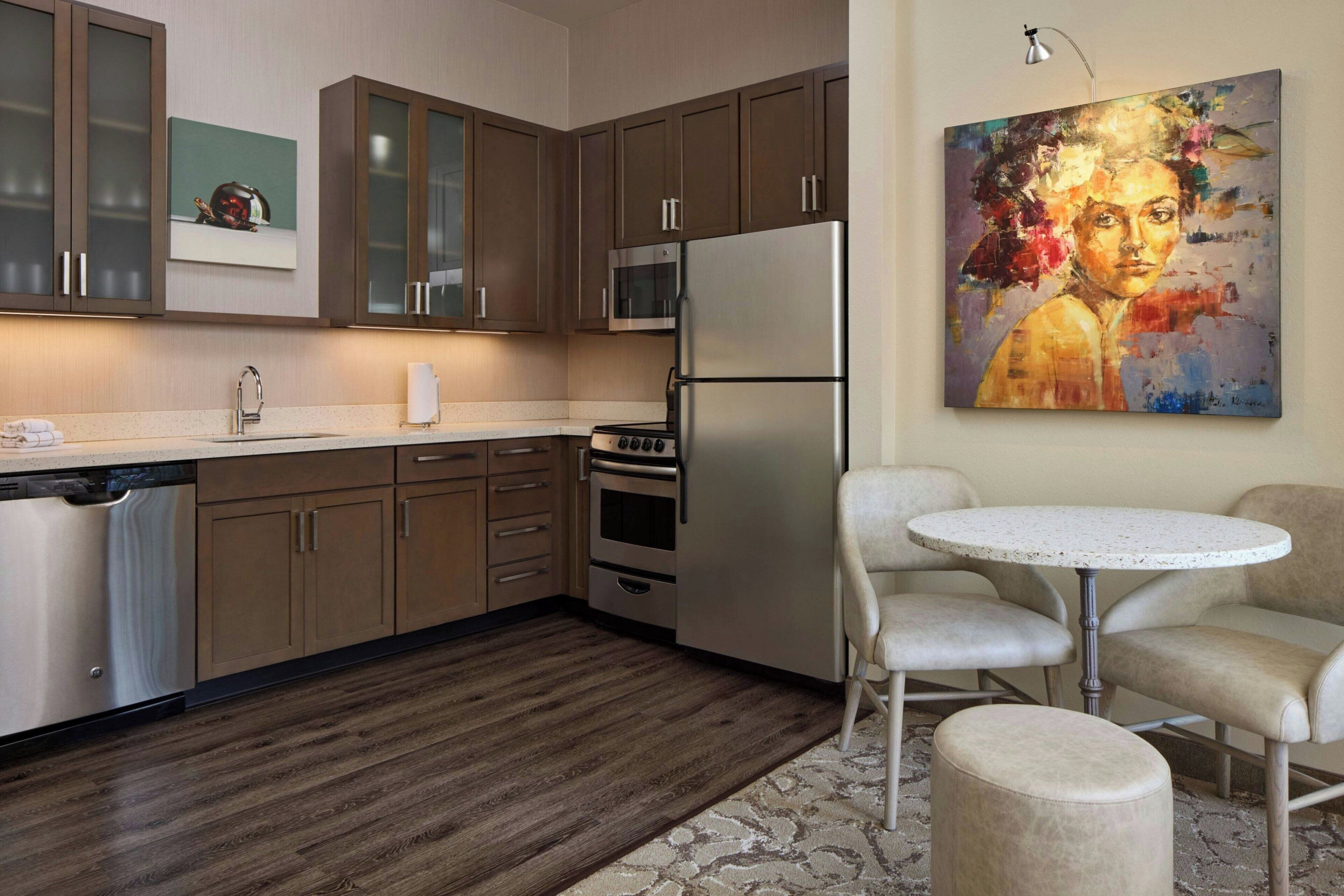 A modern kitchen with wooden cabinets, stainless steel appliances, a dining area with a round table and chairs, and a painting on the wall.