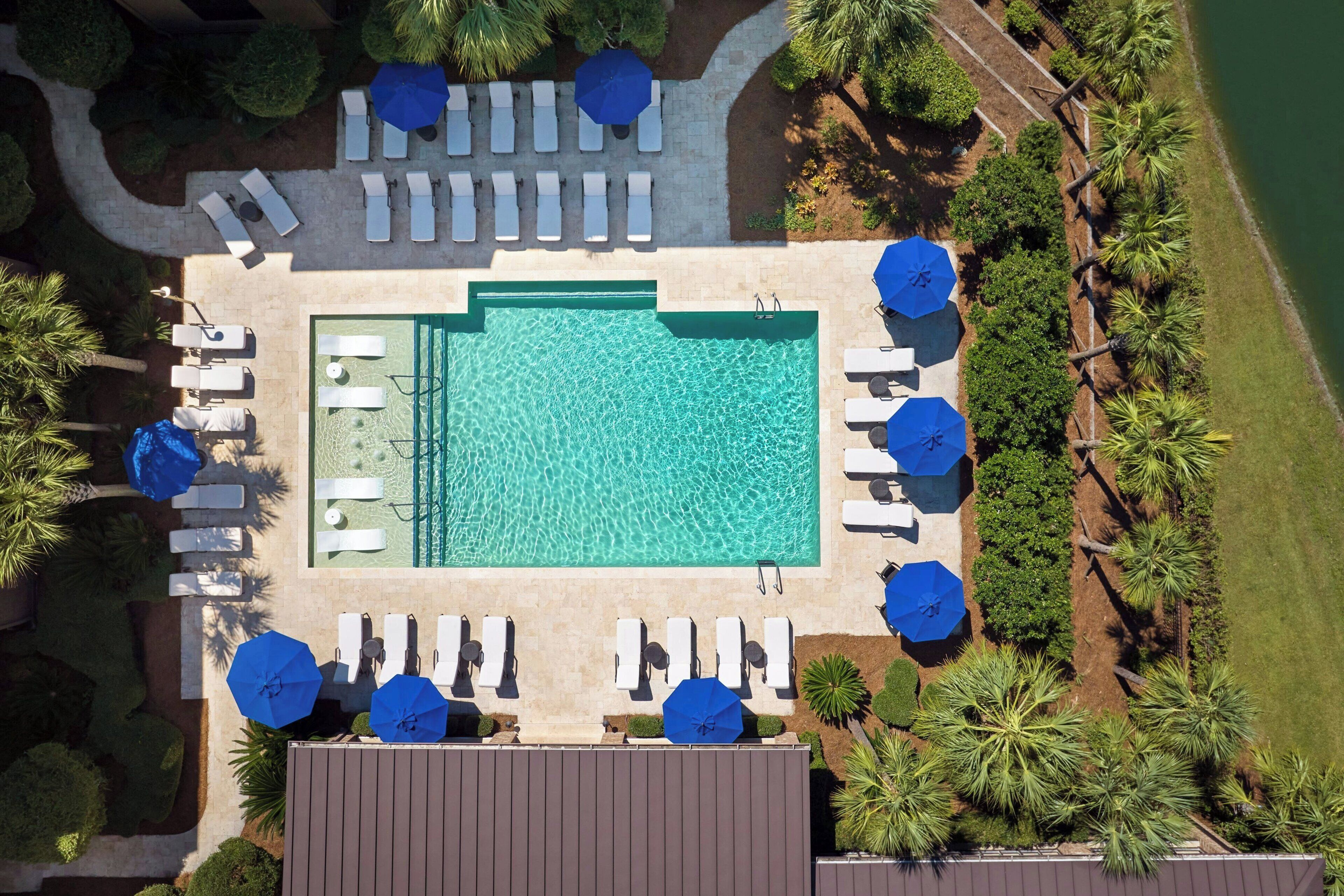 An aerial view of a pool area with sun loungers and umbrellas, surrounded by palm trees and a clear blue pool.