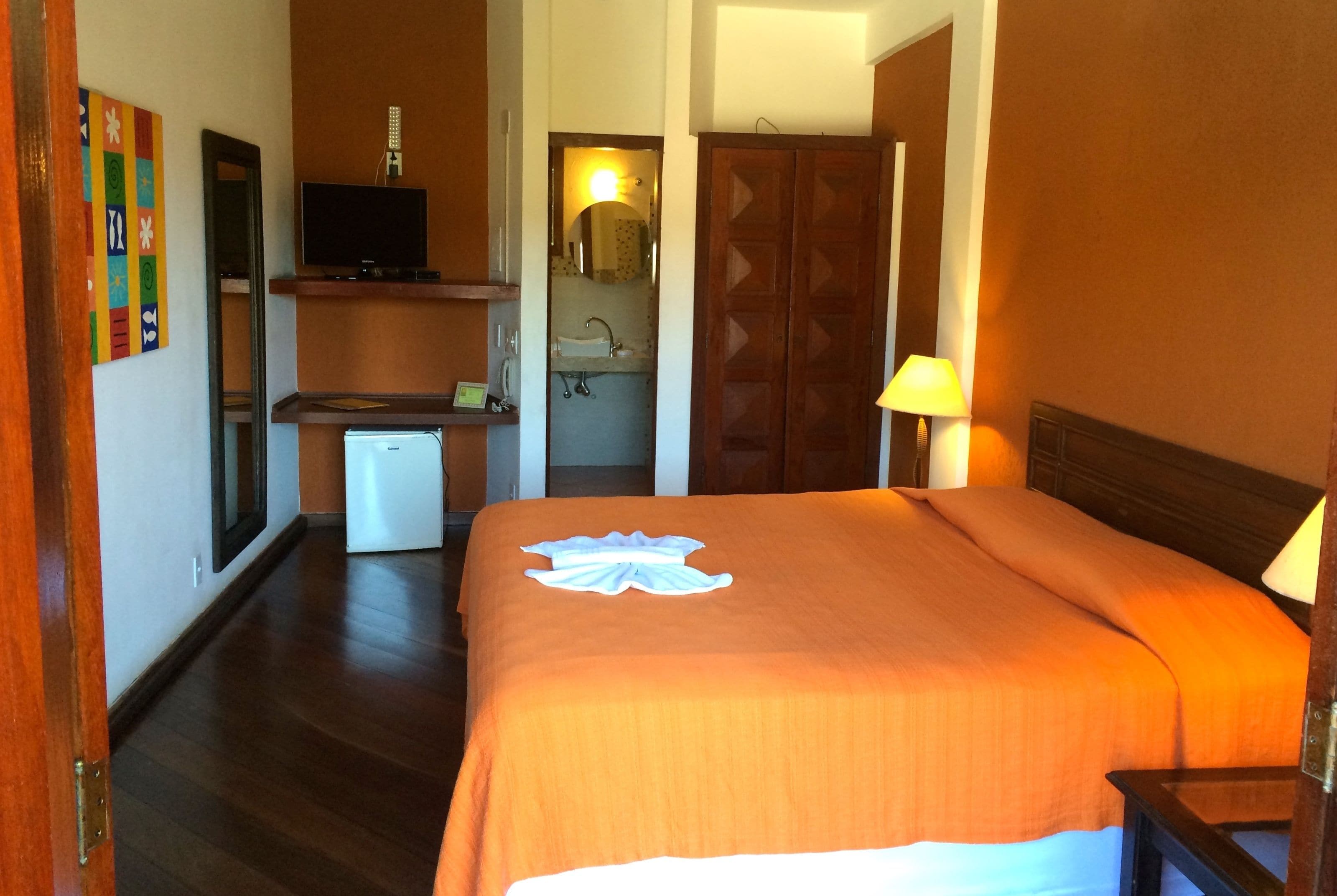 A hotel room with a bed, a desk with a television, a mirror, a bathroom with a sink, and a wooden wardrobe.