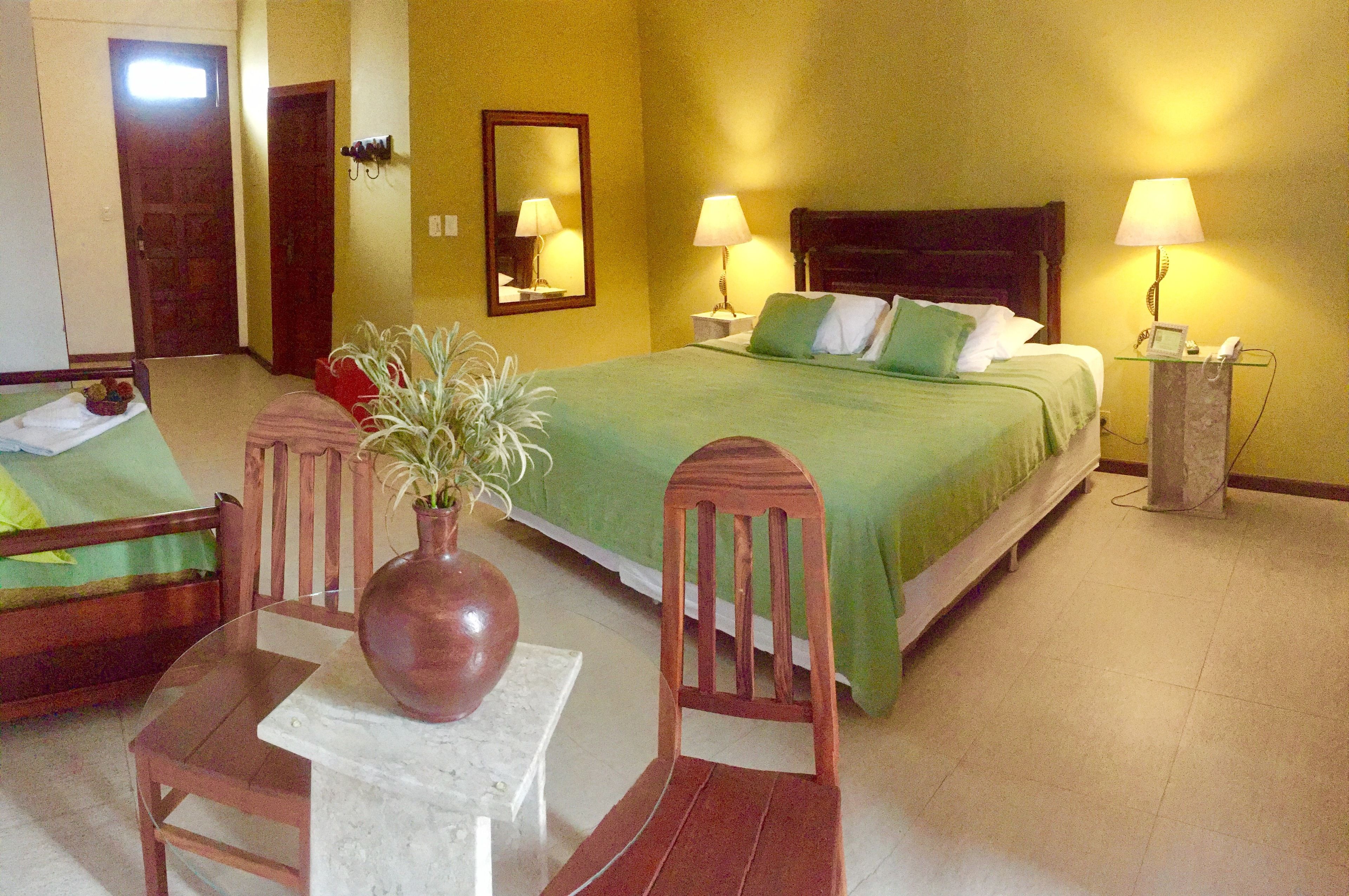 A hotel room with a bed, two bedside tables with lamps, a wooden chair, a small table with a vase, and a mirror on the wall.