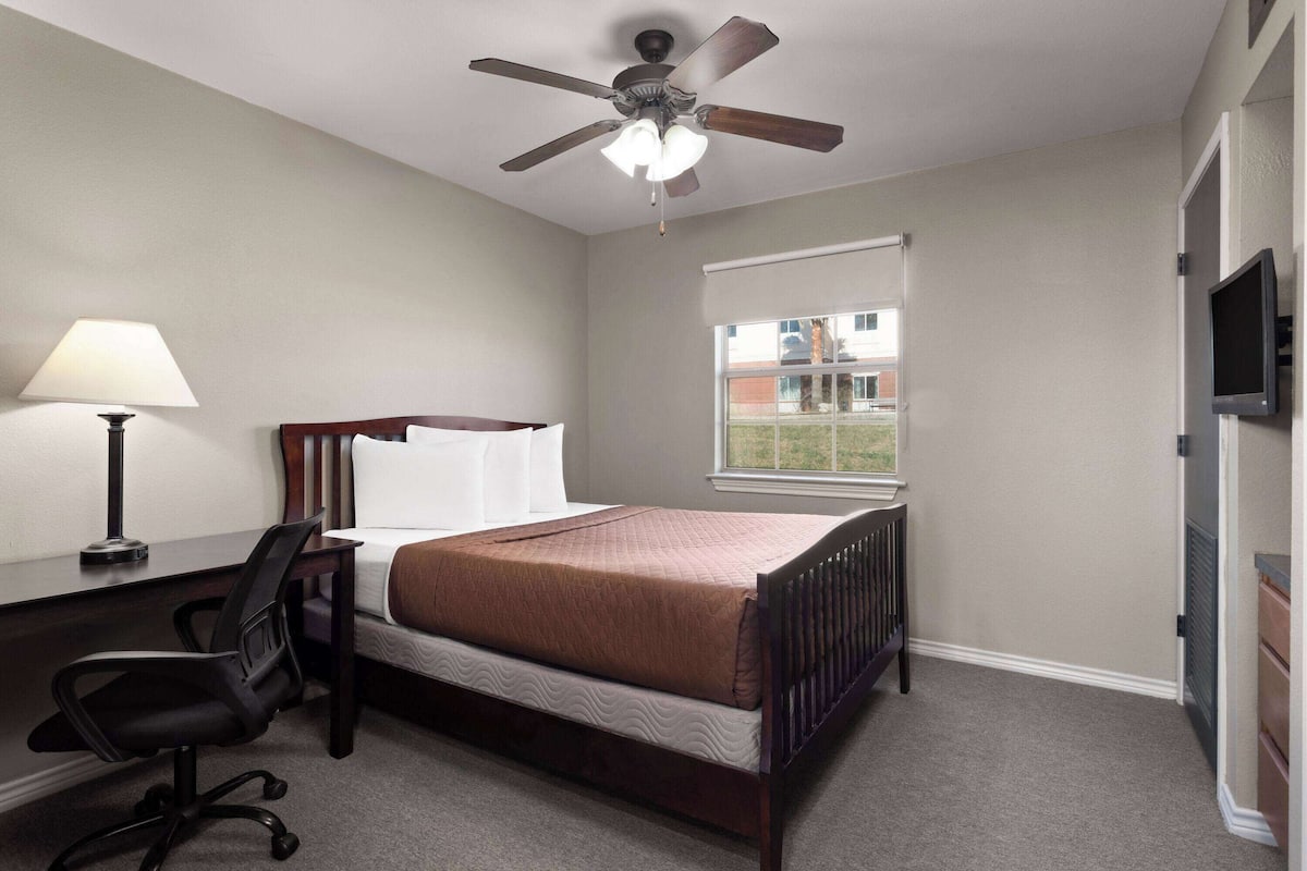 Une chambre à coucher comprenant un lit, un bureau, une chaise, un ventilateur de plafond, une fenêtre et un téléviseur.