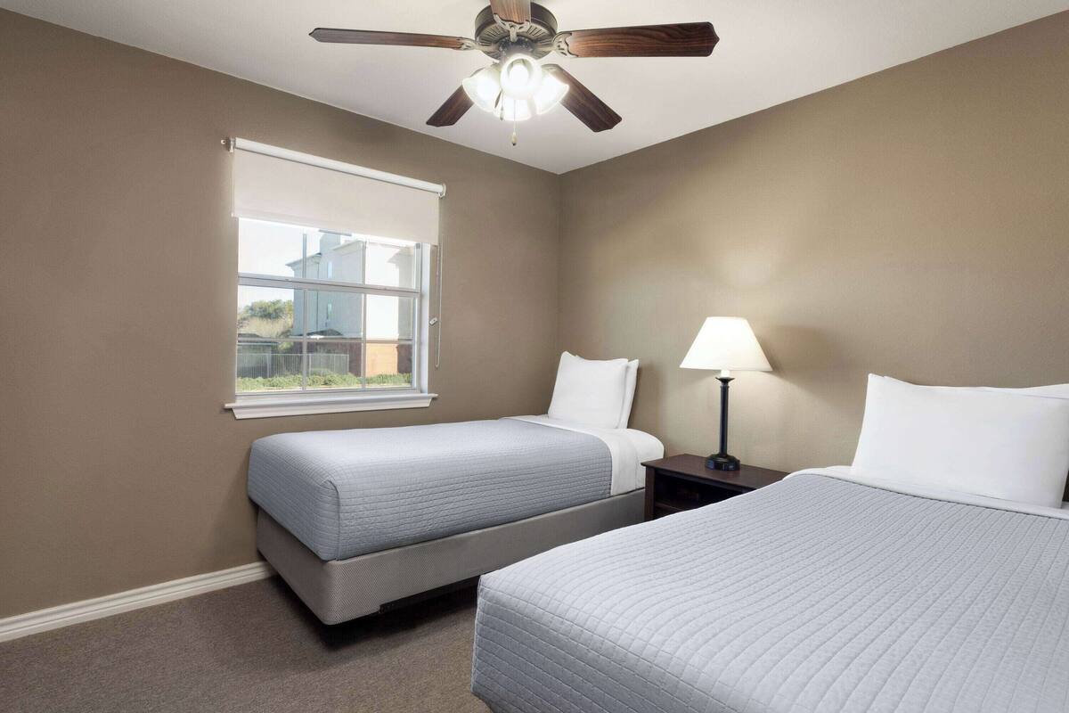 Une chambre avec deux lits, un ventilateur de plafond, une fenêtre et une table de chevet avec une lampe.