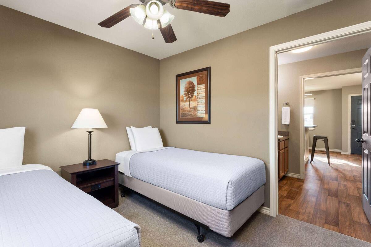 Une chambre d’hôtel avec deux lits, un ventilateur de plafond, une table de chevet avec une lampe, un tableau encadré au mur et une salle de bain visible à travers une porte ouverte.
