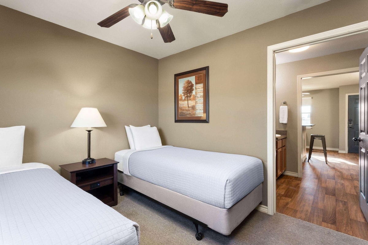A hotel room with two beds, a ceiling fan, a nightstand with a lamp, a framed picture on the wall, and a bathroom visible through an open door.