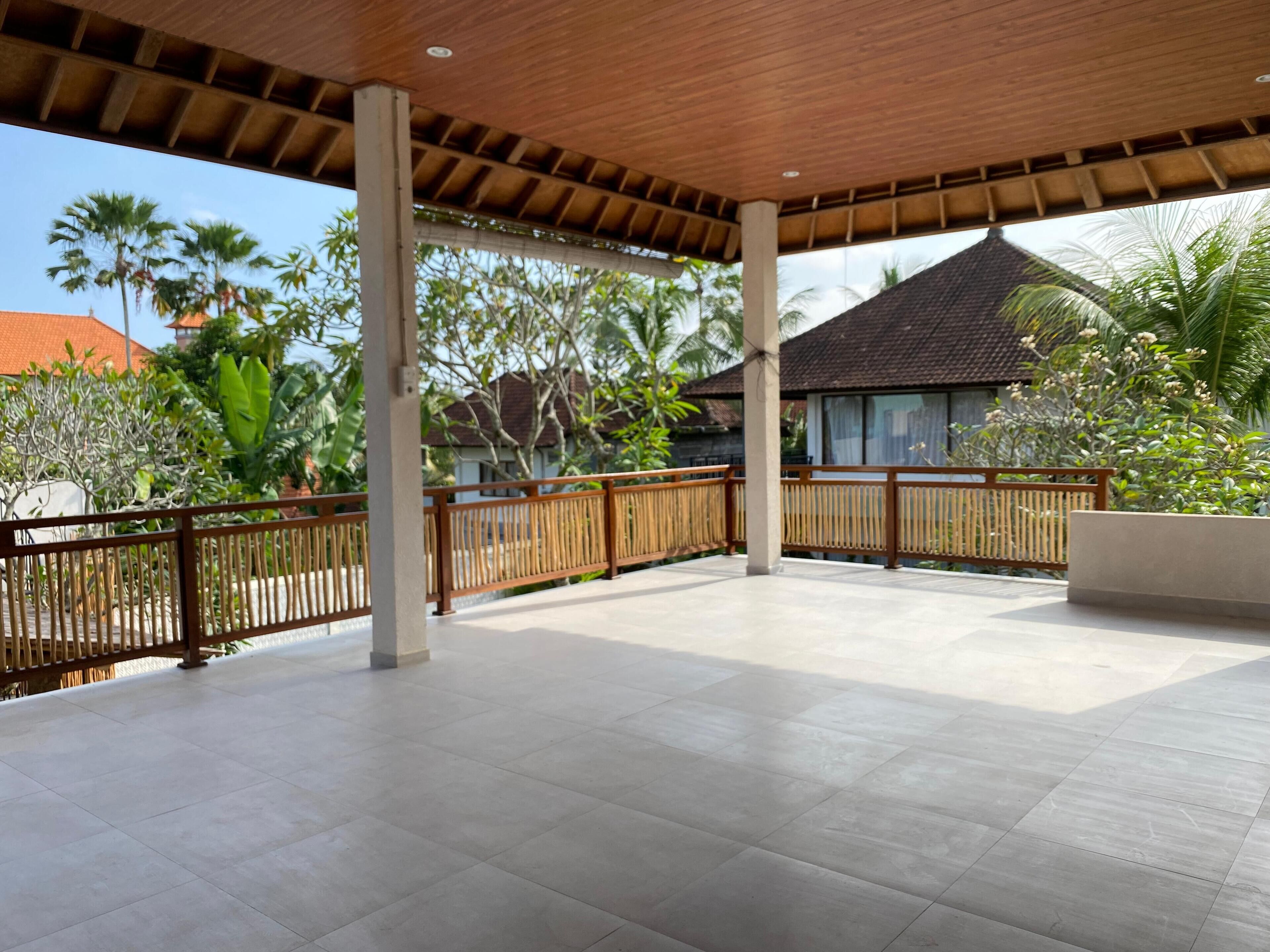 A covered outdoor area with tiled flooring, wooden railings, and a view of surrounding buildings and greenery.