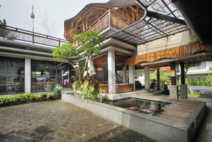 A covered outdoor area with a water feature, seating, and a building with a wooden balcony. - Putri Ayu Cottages (Ubud)