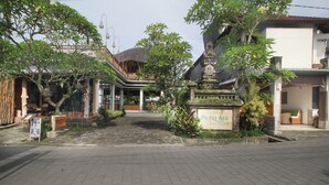 Une cour avec une enseigne indiquant « Putri Ayu Cottages », entourée d’une végétation luxuriante et bordée d’édifices à l’architecture traditionnelle.