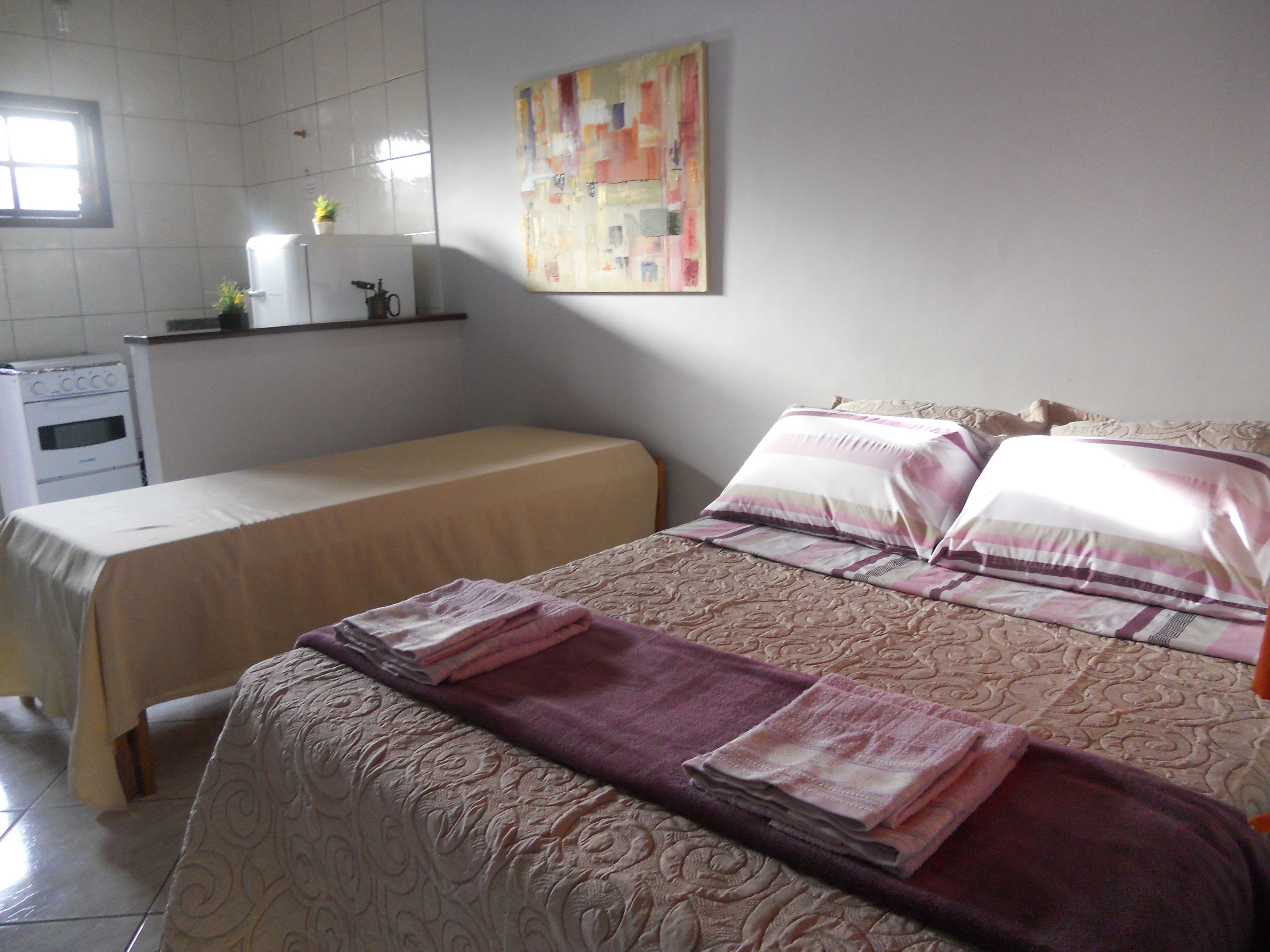 A bedroom with a bed, a bedside table, a small window, and a kitchen area with a stove and sink.