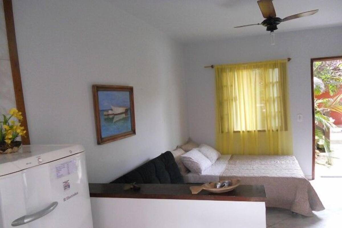 Quarto compacto com uma cama, escrivaninha, geladeira, ventilador de teto e janela com cortinas amarelas.