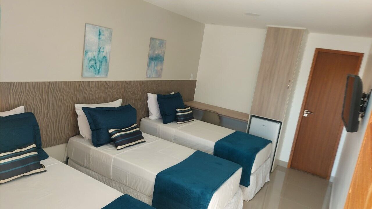 A hotel room with three single beds, each with a blue bedspread and pillows. There are two abstract paintings on the wall, a wooden headboard, and a television on a stand.
