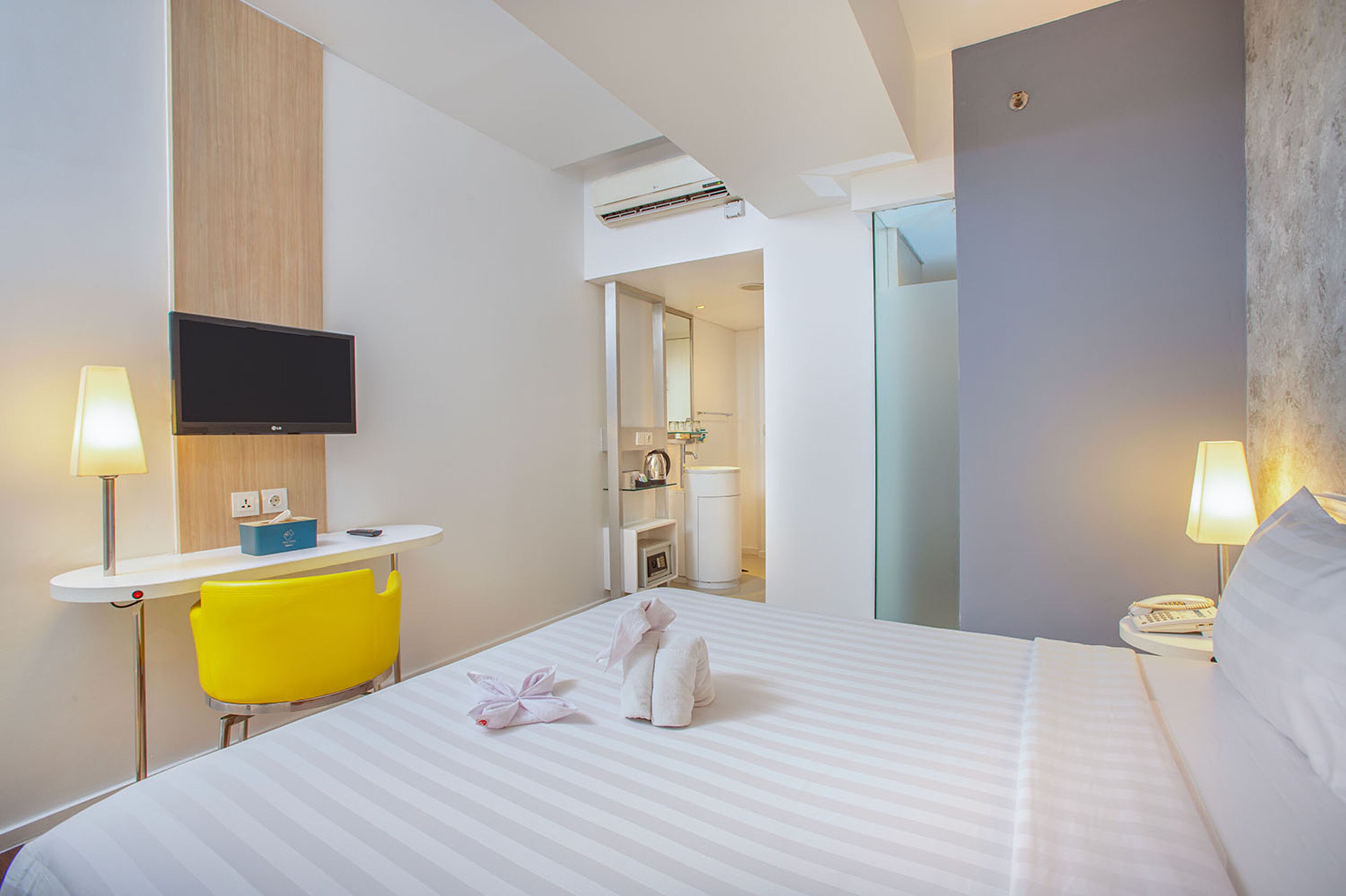 Quarto de hotel com cama, televisão, escrivaninha, cadeira e banheiro visível por uma porta aberta.