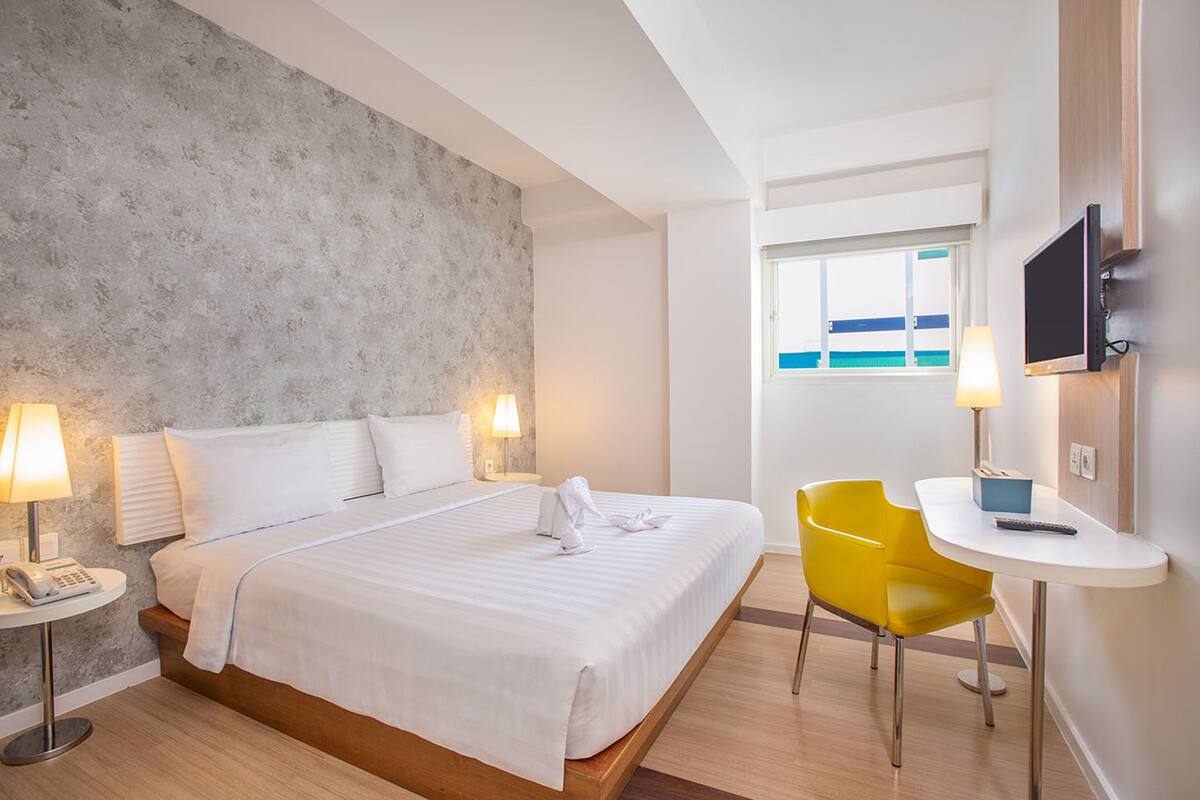 Ein Hotelzimmer mit einem Bett, Nachttischlampen, einem kleinen Tisch mit Stuhl, einem Fernseher und einem Fenster mit Meerblick.