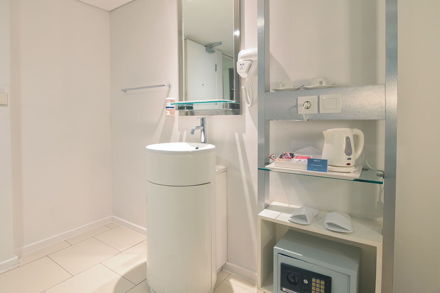 Un baño moderno con un lavamanos blanco, cabina de ducha de vidrio, espejo, hervidor eléctrico blanco y una caja fuerte pequeña.