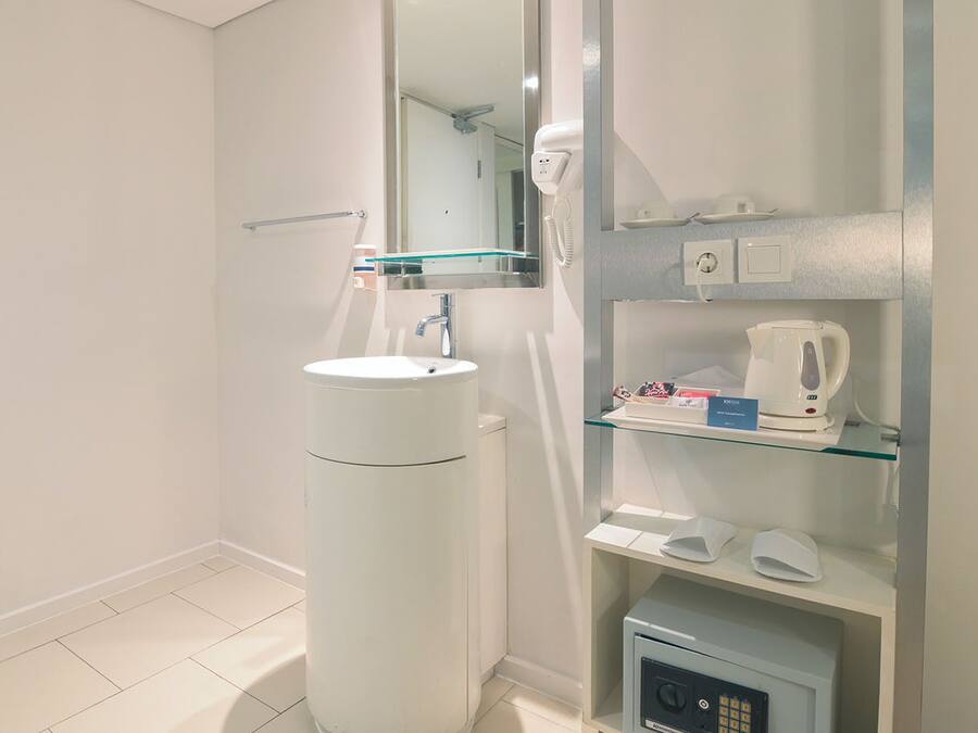 Un baño moderno con un lavamanos blanco, cabina de ducha de vidrio, espejo, hervidor eléctrico blanco y una caja fuerte pequeña.