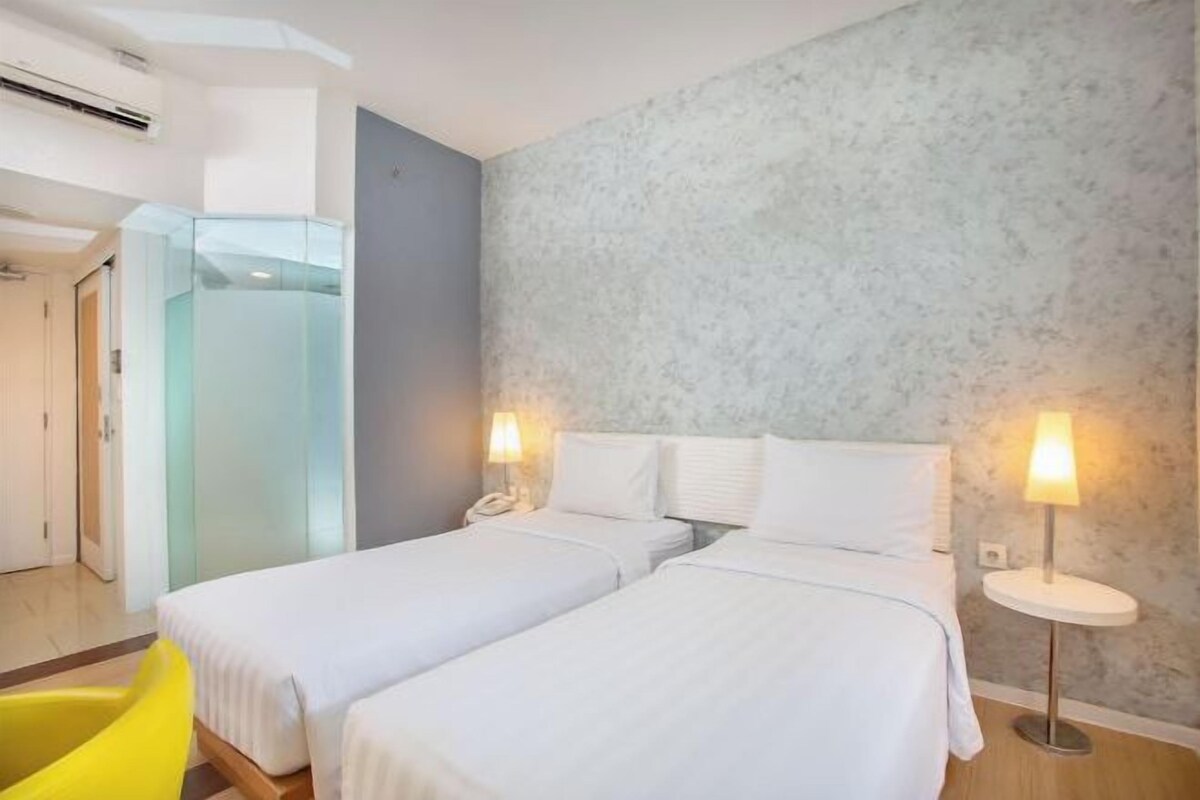 Une chambre d’hôtel avec deux lits, une cloison en verre, une chaise jaune et une lampe fixée au mur.
