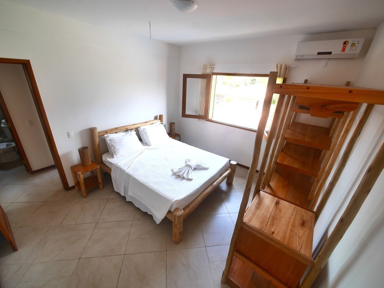 A bedroom with a wooden bed, a small wooden chair, a wooden staircase, and a window with curtains.