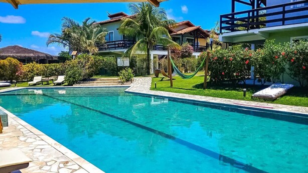 A swimming pool with a clear blue water, surrounded by a well-maintained lawn, palm trees, and a hammock. - Village Mutà (Porto Seguro)