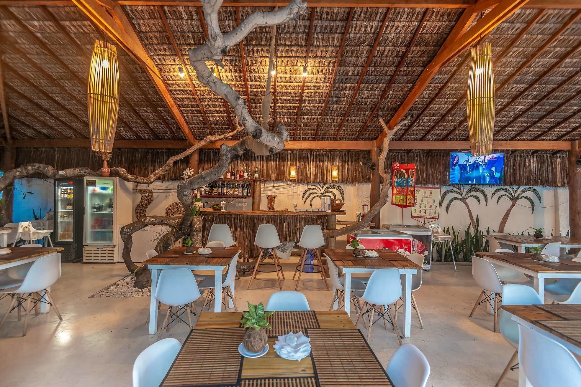 A restaurant with a rustic interior, featuring a thatched roof, wooden tables and chairs, and a bar area with a TV screen.