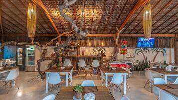A restaurant with a rustic interior, featuring a thatched roof, wooden tables and chairs, and a bar area with a TV screen.