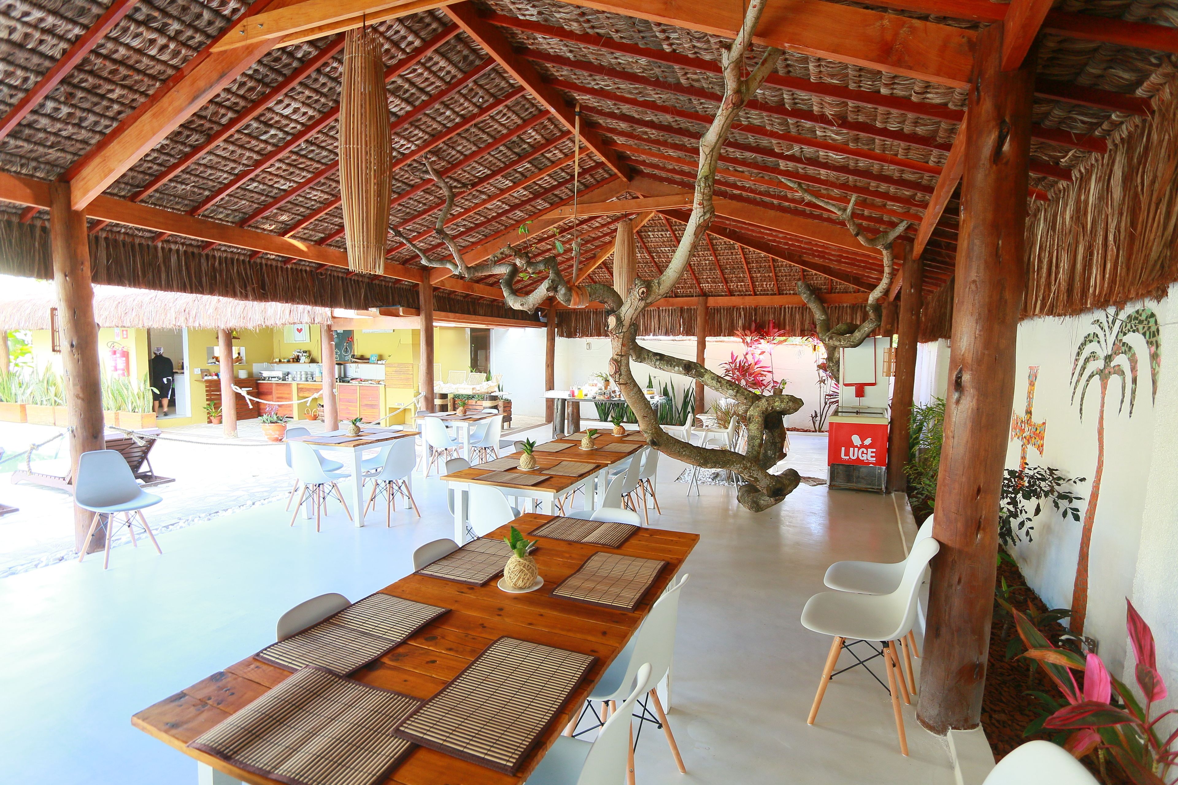 Uma sala de jantar integrada com mesas de madeira e cadeiras brancas, teto de palha e vista para a área externa.