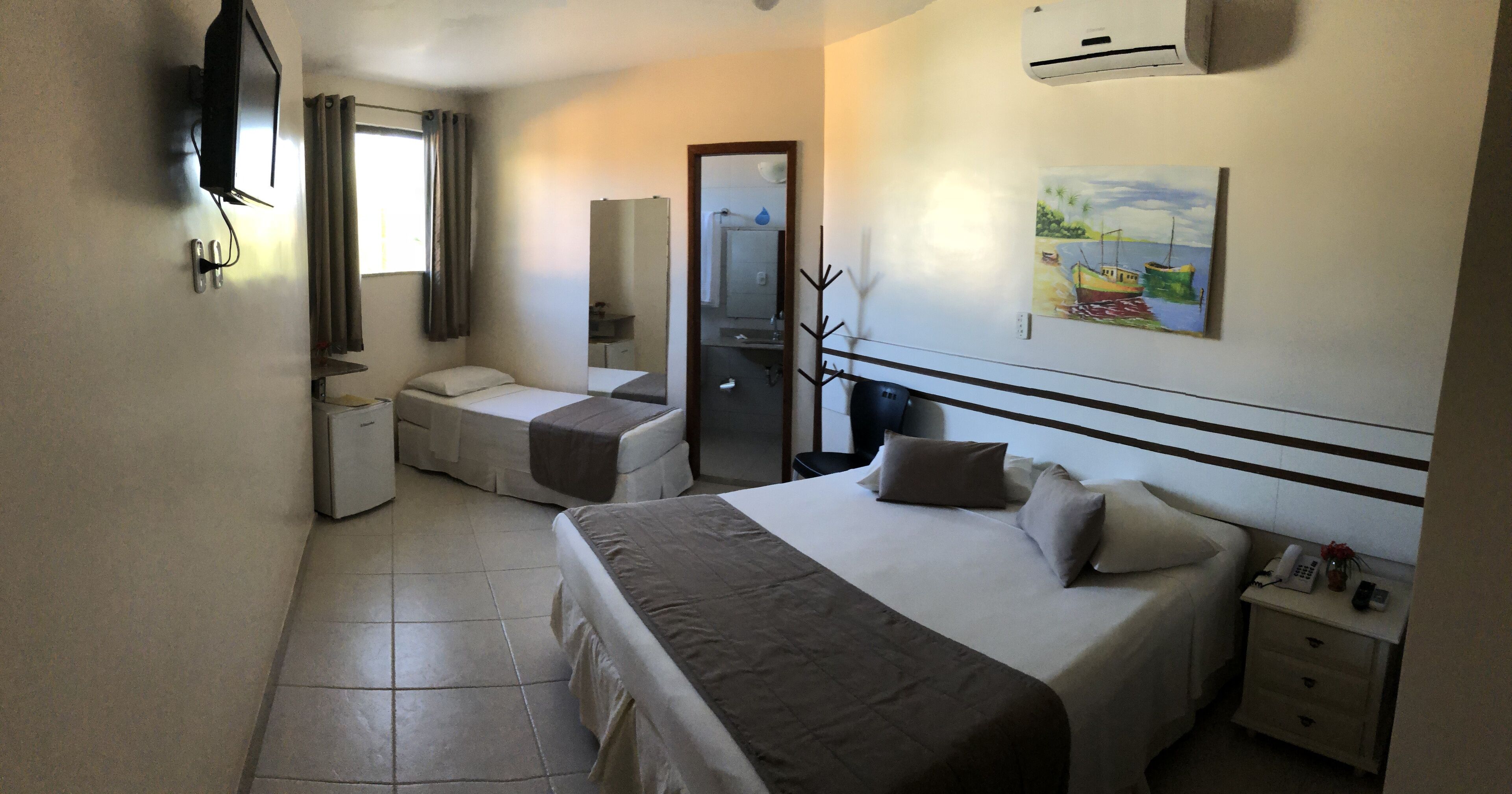 A hotel room with a bed, a smaller bed, a desk with a phone, a mirror, and a painting on the wall.