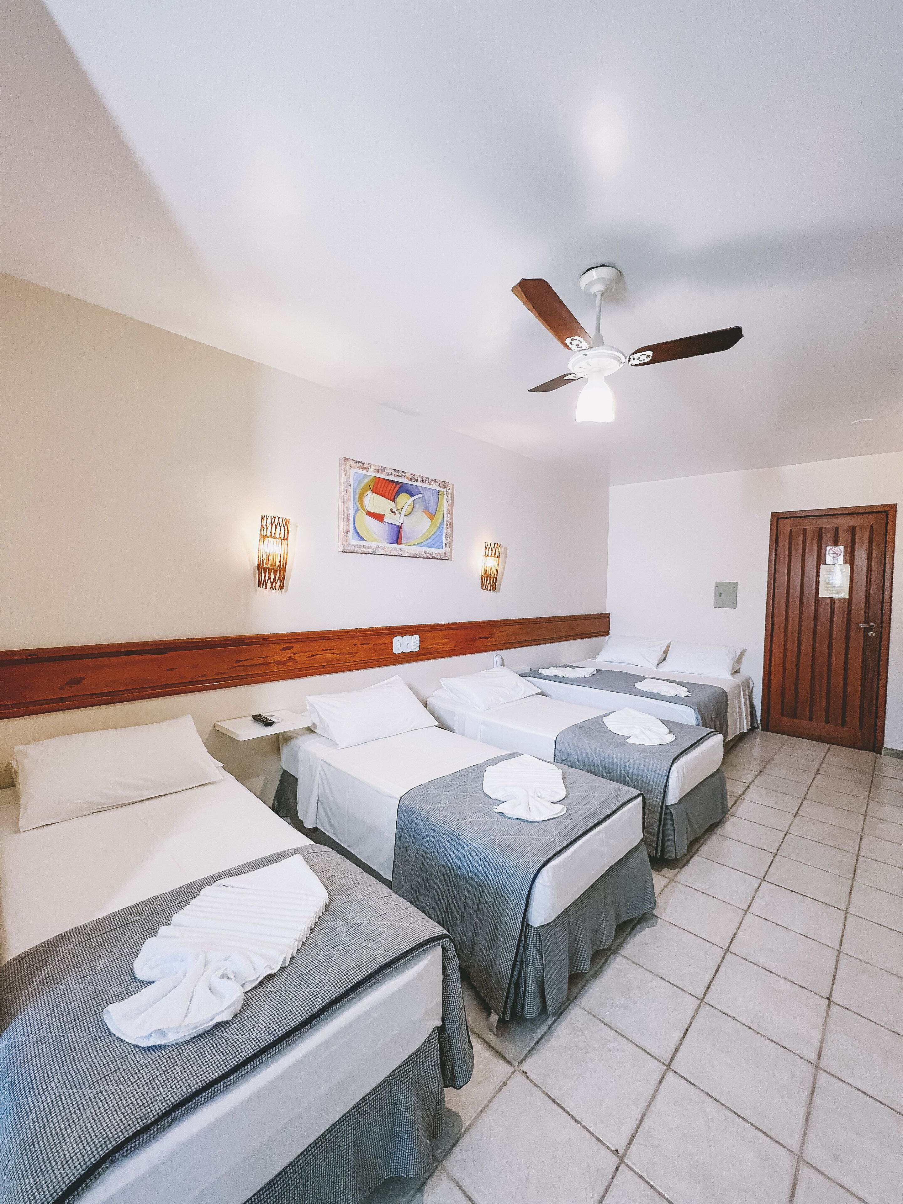 A hotel room with three beds, a ceiling fan, wall-mounted lights, and a door.